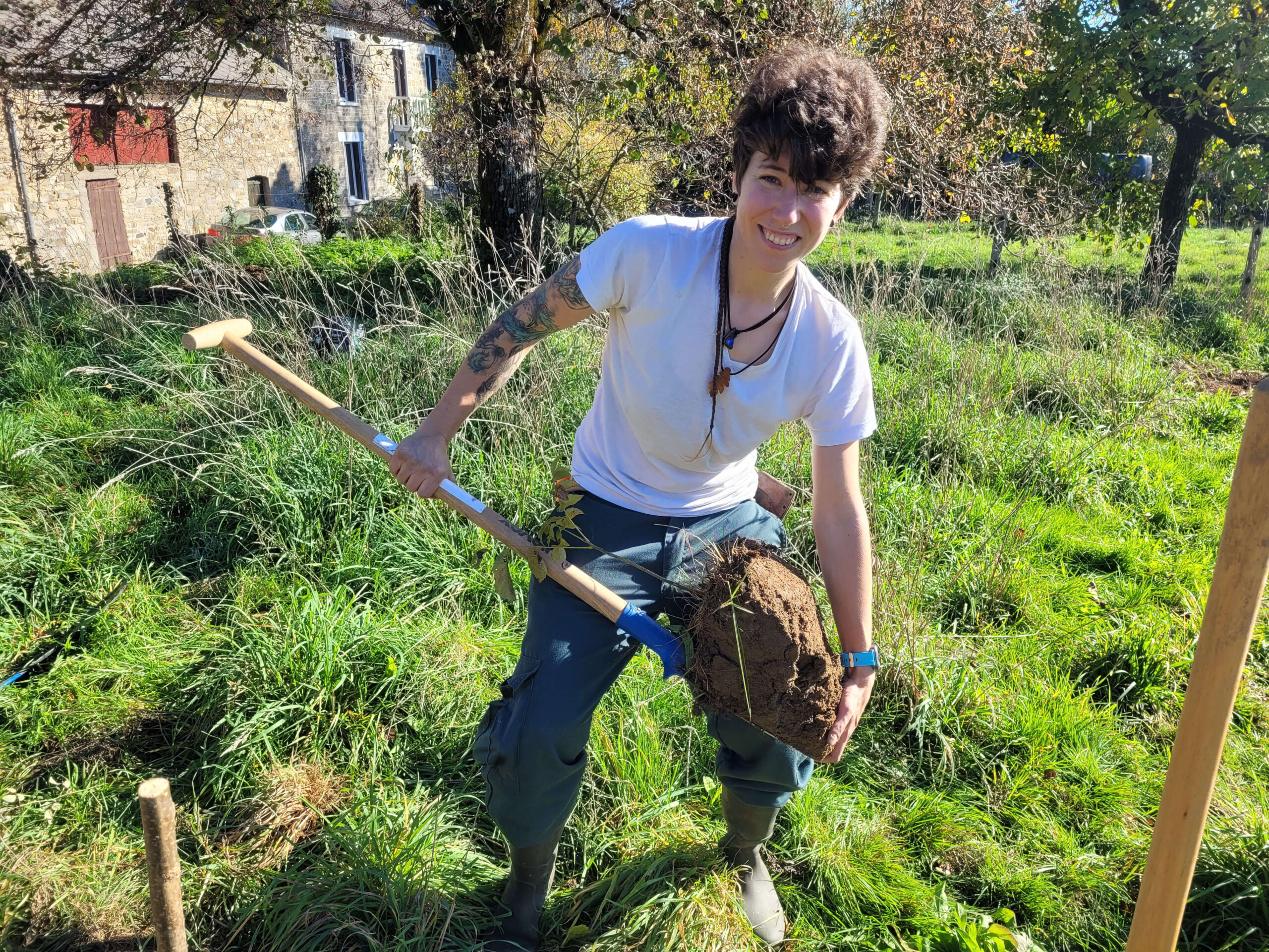 Mathilde souriante, transplantant un jeune noyer spontané avec une motte de terre généreuse, tenant une bêche dans notre jardin forêt en permaculture.